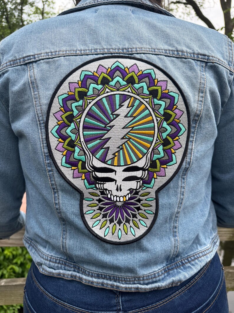 Pode incluir: Casaco de ganga azul claro com um grande patch colorido nas costas. O patch apresenta uma caveira estilizada com um raio e um design de mandala em tons de roxo, verde-azulado, amarelo e verde.