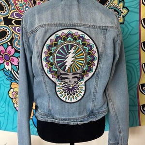Pode incluir: Casaco de ganga azul claro com um grande patch colorido nas costas. O patch apresenta uma caveira, um raio e um design floral em tons de roxo, verde e amarelo. O casaco tem uma gola cl&aacute;ssica e fechos de bot&atilde;o.