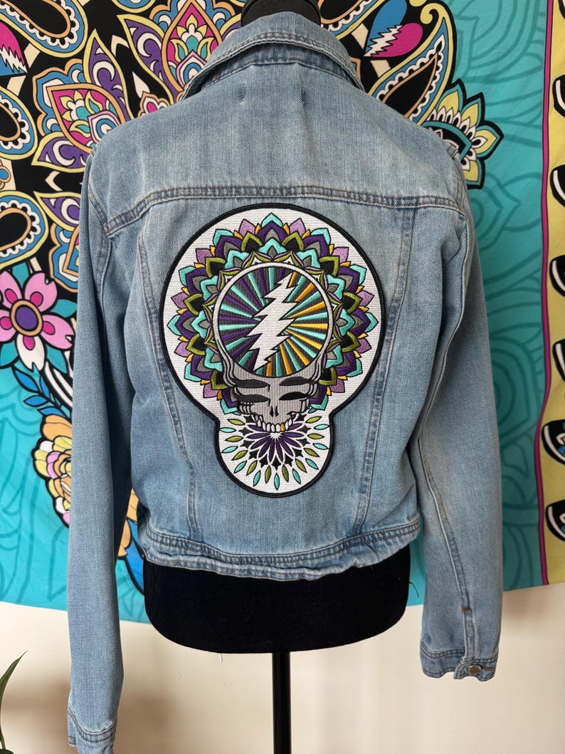 Pode incluir: Casaco de ganga azul claro com um grande patch colorido nas costas. O patch apresenta uma caveira, um raio e um design floral em tons de roxo, verde e amarelo. O casaco tem uma gola cl&aacute;ssica e fechos de bot&atilde;o.