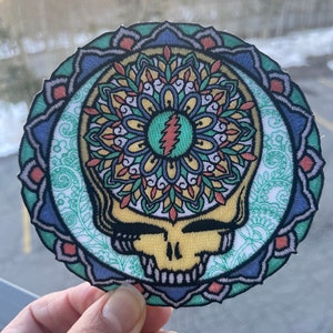 Könnte beinhalten: Ein runder gestickter Patch mit einem farbenfrohen Mandala-Design und einem Grateful Dead Steal Your Face-Schädel in der Mitte. Das Mandala ist in Blautönen, Grün, Gelb und Rot gehalten.