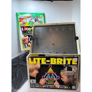 May include: A vintage Lite-Brite toy set with a black screen and a box featuring a clown design made of colorful pegs. A DuckTales picture set is visible in the background. The box has the text "LITE-BRITE" and "MAGIC TOY".
