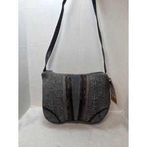 May include: Gray crossbody bag with a woven texture and vertical stripes in shades of gray, brown, and black. The bag has a black strap and a zipper closure. The bottom corners are reinforced with gray suede.