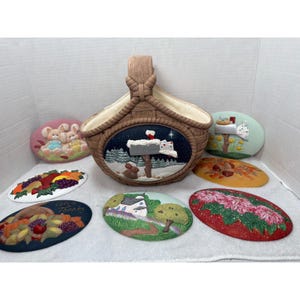 May include: A brown ceramic basket with a woven design and a handle, surrounded by several oval-shaped decorative plaques. The plaques feature various seasonal scenes, including bunnies, autumn leaves, and a winter mailbox scene.