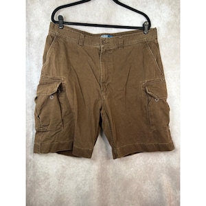 May include: Brown cargo shorts featuring a button-fly closure, belt loops, and two large side pockets with buttoned flaps. The waistband includes a small blue Polo Ralph Lauren label. These shorts are constructed with visible stitching and a structured fit.