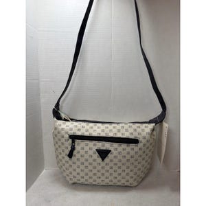 May include: Cream-colored crossbody bag with a repeating geometric pattern in navy blue. The bag features a black zipper, a black strap, and a black triangle emblem. A tag hangs from the side.