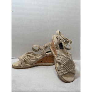 Puede incluir: Un par de sandalias de cuña beige. Las sandalias presentan correas tejidas y un tacón de cuña de corcho. Las correas del tobillo tienen un cierre de hebilla. Los zapatos están hechos de materiales naturales y tienen un estilo casual y veraniego.