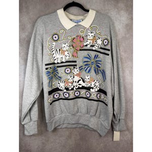 May include: A grey sweatshirt with a white collar, featuring a cat design. The design includes striped and spotted cats with bows, decorative bows, and floral details. The sweatshirt has long sleeves and a relaxed fit.