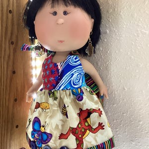 May include: A handmade doll with black hair and a painted face. The doll wears a colorful dress with a red and blue top and a beige skirt featuring a butterfly and frog print. The dress is trimmed with a striped border and adorned with large buttons.