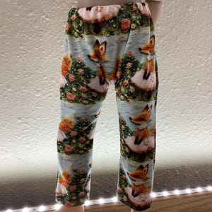 May include: A pair of patterned leggings featuring a whimsical design. The leggings display a repeating pattern of foxes in a garden setting with flowers and a light blue background. The color palette includes shades of orange, pink, and green.