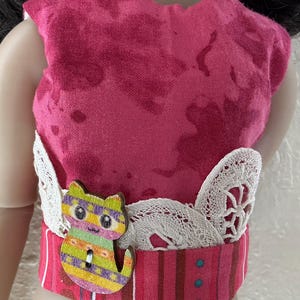 May include: A doll's pink top with a decorative striped belt and a wooden cat-shaped button. The top is made of pink fabric with darker pink splotches. The belt has white lace accents.