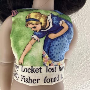 Puede incluir: Una camiseta para muñeca con una colorida ilustración de una niña con un vestido azul sobre un fondo verde. El texto "Lucy Locket lost her Kitty Fisher found it" está impreso en la parte inferior.