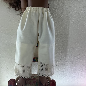 May include: Cream-colored doll pants with a crochet lace trim at the hem. The pants have a loose, comfortable fit and a drawstring waist. The doll pants are made of a lightweight fabric.