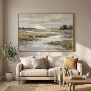 May include: A framed landscape painting shows a marsh scene with a white bird. The artwork uses muted tones of beige, green, and blue, with a cloudy sky. The painting is displayed above a beige sofa with cushions and a wooden coffee table.