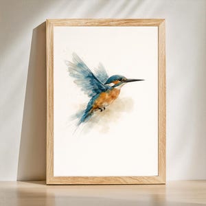Op de afbeelding: Ingelijste aquarel van een ijsvogel in vlucht. De vogel heeft blauwe vleugels, een oranje borst en een lange zwarte snavel. Het kunstwerk wordt gepresenteerd in een licht houten frame, met een zachte, artistieke stijl.