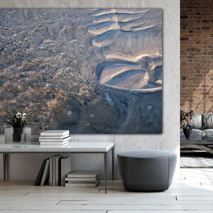 Może przedstawiać: Duży wydruk na płótnie przedstawiający widok z lotu ptaka na piaszczystą plażę z wodą. Grafika zawiera odcienie niebieskiego, beżowego i brązowego, z widocznymi fakturami piasku i wody. Wydruk jest wyświetlany nad białym stołem z książkami i małą rośliną.