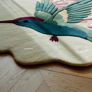 K&ouml;nnte beinhalten: Handgefertigter Teppich mit einem farbenfrohen Kolibri-Design. Der Vogel hat t&uuml;rkisfarbene, gr&uuml;ne und violette Akzente mit rosa Blumendetails. Der Teppich ist cremefarben und liegt auf einem Holzboden. Der Text "RugHandmade" ist sichtbar.