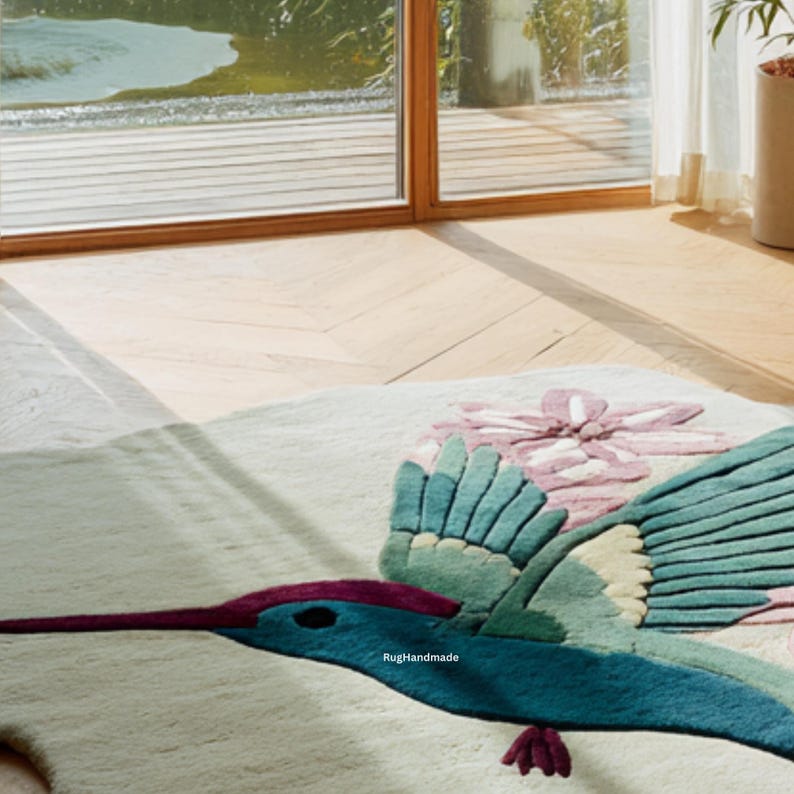 K&ouml;nnte beinhalten: Ein handgefertigter Teppich mit einem Kolibri-Design. Der Teppich hat einen hellen Hintergrund mit einem gro&szlig;en Kolibri in T&uuml;rkis-, Blau- und Lilat&ouml;nen. Eine rosa Blume ist ebenfalls auf dem Teppich abgebildet. Der Text "RugHandmade" ist sichtbar.