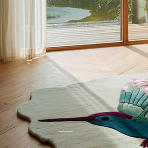 K&ouml;nnte beinhalten: Ein handgefertigter, cremefarbener Teppich mit einem Kolibri-Design. Der Teppich zeigt einen t&uuml;rkisblauen Kolibri mit einem langen, lila Schnabel und gr&uuml;nen Fl&uuml;geln. Das Design umfasst rosa und gr&uuml;ne florale Akzente. Der Teppich liegt auf einem Hartholzboden.