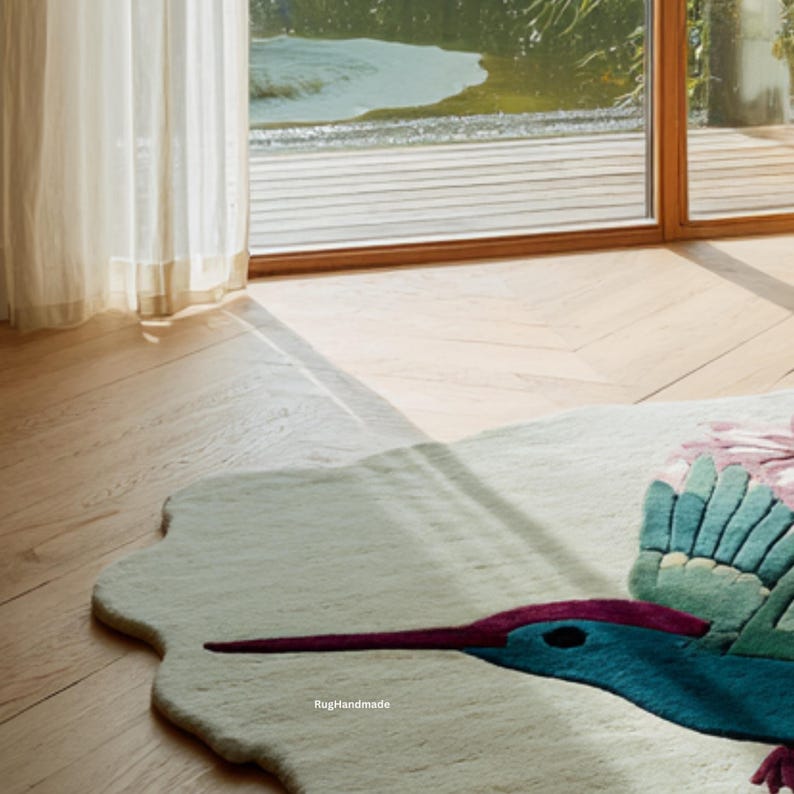 K&ouml;nnte beinhalten: Ein handgefertigter, cremefarbener Teppich mit einem Kolibri-Design. Der Teppich zeigt einen t&uuml;rkisblauen Kolibri mit einem langen, lila Schnabel und gr&uuml;nen Fl&uuml;geln. Das Design umfasst rosa und gr&uuml;ne florale Akzente. Der Teppich liegt auf einem Hartholzboden.