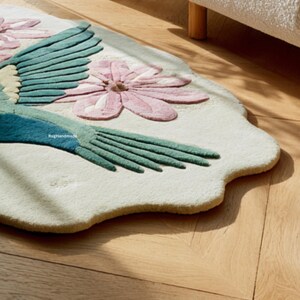 K&ouml;nnte beinhalten: Ein cremefarbener, einzigartig geformter Teppich mit einem Vogel- und Blumenmuster. Der Vogel hat t&uuml;rkisfarbene und blaue Fl&uuml;gel und Schwanzfedern, mit einer rosa Blume in der N&auml;he. Der Text "Rug Handmade" ist sichtbar.