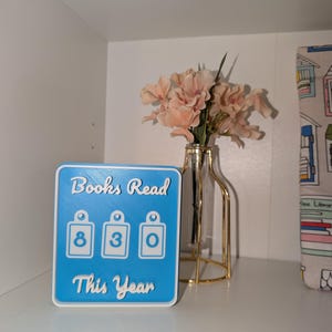 May include: A blue and white "Books Read This Year" counter with the numbers 830 displayed. The counter is on a shelf with a vase of pink flowers and a decorative item with a house pattern.