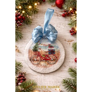 May include: A round Christmas ornament with a light blue satin bow at the top. The ornament features a red bicycle carrying a Christmas tree and gifts. The ornament is surrounded by a white, fluffy border.