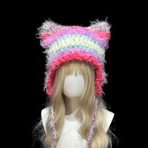 以下が含まれることがあります： カラフルな手作りの猫耳帽子。ピンク、紫、黄色、青など、虹色の色合いが特徴です。長い三つ編みの紐が付いており、ブロンドの髪のマネキンの頭に飾られています。