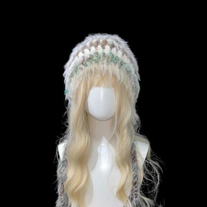 May include: A fluffy, handmade hat with a light blonde wig attached. The hat features a mix of white, brown, and green yarn, with long, braided strands hanging down. The wig has long, wavy hair.