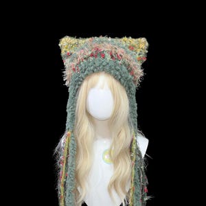 Puede incluir: Un sombrero hecho a mano con forma de orejas de gato, de color verde, con largas franjas colgantes. El sombrero está hecho de un hilo difuso y texturizado con acentos rosas, amarillos y rojos. El sombrero se muestra en una cabeza de maniquí con pelo largo y rubio.