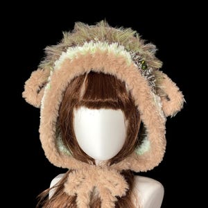 May include: A fluffy, hooded hat in brown, beige, and green, with a faux fur texture. The hat has small ear-like shapes and a bow at the neck. The hat is designed to be worn on the head.
