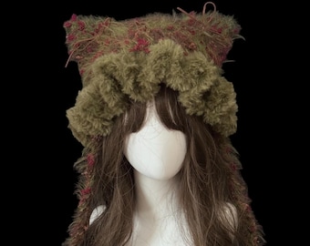 May include: A whimsical, handmade hat with a unique design. The hat features a combination of olive green and burgundy accents, with a fluffy texture and ear-like protrusions. The hat is displayed on a mannequin head with long brown hair.