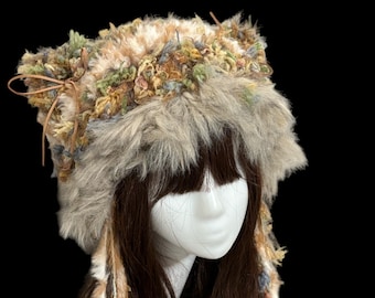 Op de afbeelding: Een unieke, handgemaakte hoed met een imitatiebont afwerking en decoratieve accenten. De hoed heeft een mix van texturen en kleuren, waaronder bruin, beige, groen en blauw. De hoed heeft lange, bungelende stofstrengen en een klein strikje.