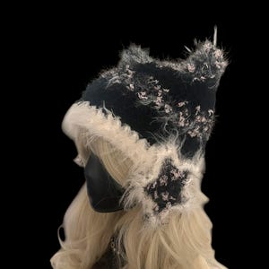 Op de afbeelding: Een zwart-witte hoed met een pluizige rand en accenten. De hoed heeft een uniek ontwerp met een textuur en staat tegen een zwarte achtergrond. De hoed is waarschijnlijk een modeaccessoire.