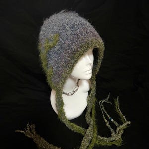 May include: A handmade, hooded hat in shades of gray, blue, and green. The hat has long, fringed ties and is displayed on a white mannequin head. The hat's texture appears soft and fuzzy, with a unique, artistic design.
