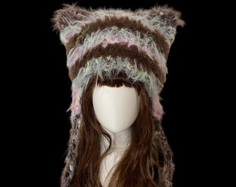 May include: A handmade, furry hat with cat ears. The hat features brown, pink, and light blue stripes. The hat is on a mannequin head with long, brown hair. The background is black.