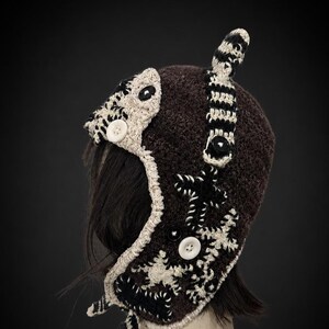 May include: A brown crocheted hat with a unique design. The hat features black and white accents, including buttons and decorative patterns. The hat has a textured appearance and a distinctive shape, suitable for cold weather.