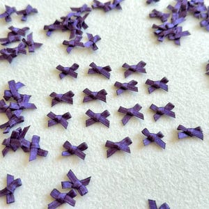 May include: A collection of small, purple satin ribbon bows scattered across the frame. Each bow is constructed from a narrow strip of fabric tied into a simple knot with two short loops and two short tails, featuring a smooth, reflective texture.