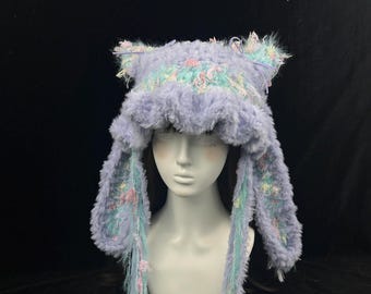 May include: A lavender faux fur hat featuring cat-like ears and long side flaps. The hat is accented with light blue and pink textured yarn details, including hanging fringe elements that extend from the sides, creating a soft, fuzzy appearance.