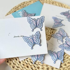 Puede incluir: Dos parches bordados de mariposa con detalles de hilo azul claro y rosa suave fijados en una tarjeta blanca. La tarjeta presenta el texto impreso "Lovely day" en tinta negra. Los parches tienen patrones de alas intrincados y bordes festoneados.