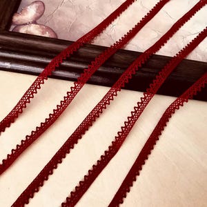 Puede incluir: Cuatro tiras paralelas de cinta picot de color rojo oscuro, cada una con un borde delicado y festoneado en ambos lados. El material estrecho y tejido muestra un tono carmesí uniforme y un patrón de zigzag repetitivo en sus bordes.