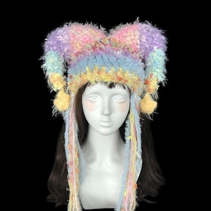May include: A colorful, fluffy hat with a jester-like design. The hat features a mix of pastel colors, including pink, purple, blue, and yellow. It has long, dangling strands and small yellow pom-poms. The hat is designed to be worn on the head.