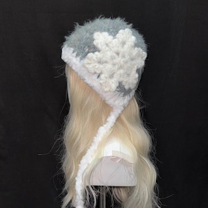 Pode incluir: Um chapéu de inverno com um design de floco de neve. O chapéu é azul claro e branco, com um grande floco de neve branco na frente. Possui protetores de ouvido e laços, e é feito de um material macio e fofo.