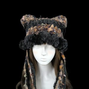 Puede incluir: Un sombrero de piel sintética con protuberancias en forma de orejas y largos flecos. El sombrero presenta una mezcla de colores negro, marrón y beige, con elementos decorativos en el fleco. El sombrero se muestra sobre una cabeza de maniquí sobre un fondo negro.