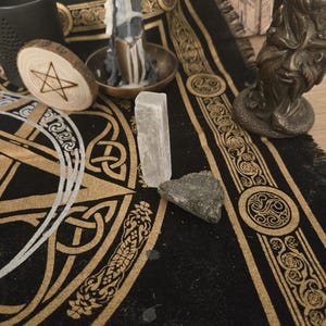 Può includere: Una collezione di oggetti spirituali su un tappetino nero e oro. Include un pentagramma di legno, una candela, un cristallo di selenite, una pietra e un portacandele color bronzo. L'immagine mostra anche una tazza nera e una statua decorativa.