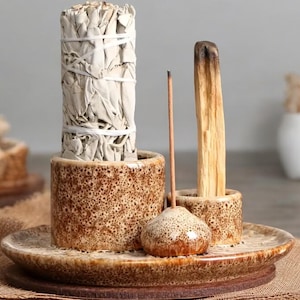 Puede incluir: Un kit de limpieza de cerámica con un manojo de salvia blanca, un palo santo, una varilla de incienso y un portaincienso decorativo. El kit se exhibe en un plato de cerámica con una base de madera marrón.