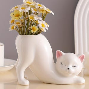 Könnte beinhalten: Eine weiße, katzenförmige Keramikvase mit gelben und weißen Blumen. Die Katze befindet sich in einer Dehnpose mit geschlossenen Augen und rosa Akzenten an den Ohren. Ein dekorativer Artikel für die Inneneinrichtung.