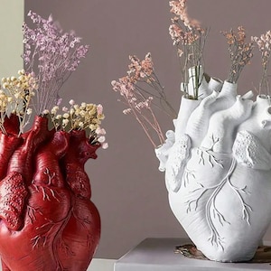 Puede incluir: Dos jarrones en forma de corazón, uno rojo y otro blanco, cada uno lleno de flores secas. Los jarrones tienen detalles anatómicos. El jarrón rojo está a la izquierda y el blanco a la derecha. El fondo es de color neutro.