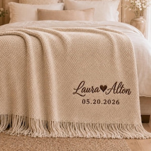 May include: A cream-coloured knit throw blanket with fringe trim, featuring dark brown embroidered text that reads Laura heart Alton and the date 05.20.2026. The blanket has a textured popcorn stitch pattern and is draped over a bed with white pillows.