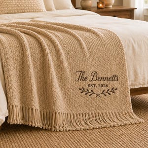 May include: A beige knitted throw blanket with fringe trim, featuring dark brown embroidery that reads The Bennetts EST. 2026 above a decorative leaf branch design. The textured blanket is draped over a bed with matching pillows.
