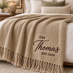 May include: A beige knit throw blanket featuring a textured diagonal pattern and long fringe along the bottom edge. The blanket is embroidered with the dark brown text "THE Thomas EST.2026" in a combination of block and script lettering.